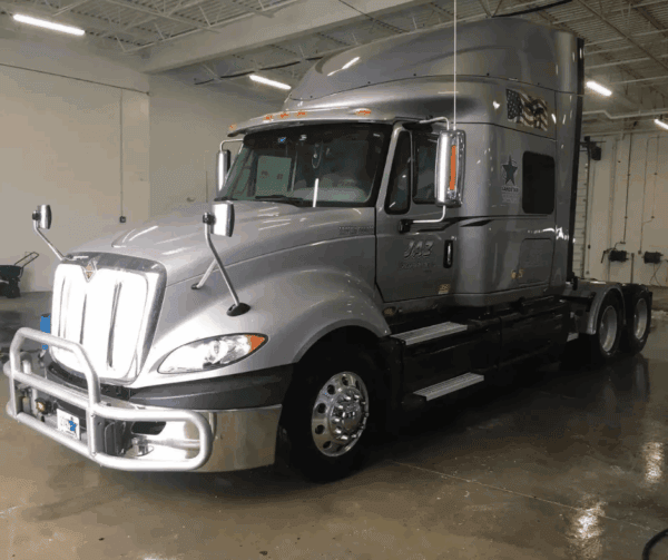 A silver semi-truck with a large cab parked indoors on a shiny, wet floor under bright overhead lights. The truck has a front grill guard and side mirrors extended.