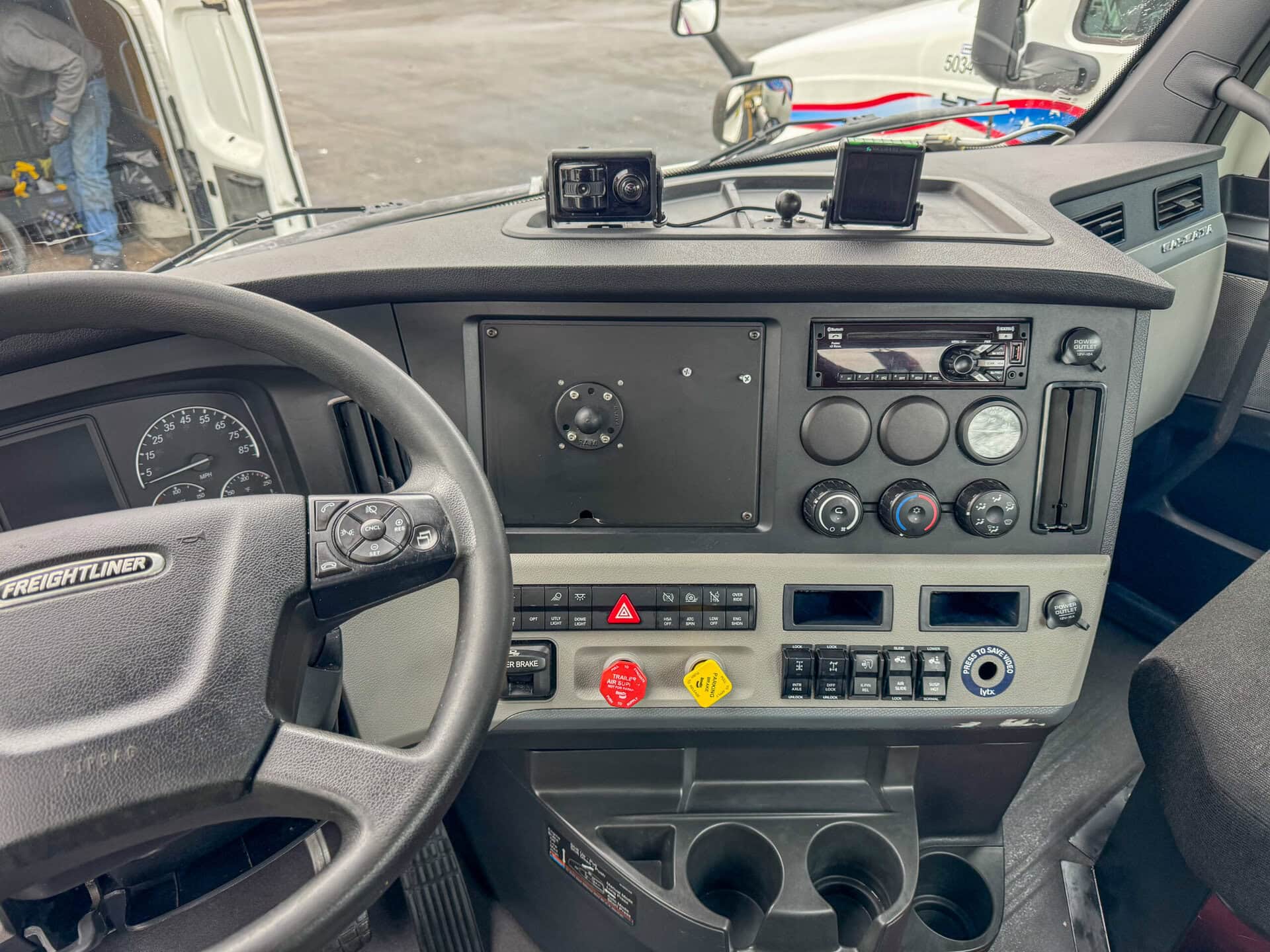 The interior cab of a Freightliner truck, showing the steering wheel, dashboard with gauges and controls, cup holders, and electronic devices mounted on the dash. Another truck and a person are visible outside through the windshield.