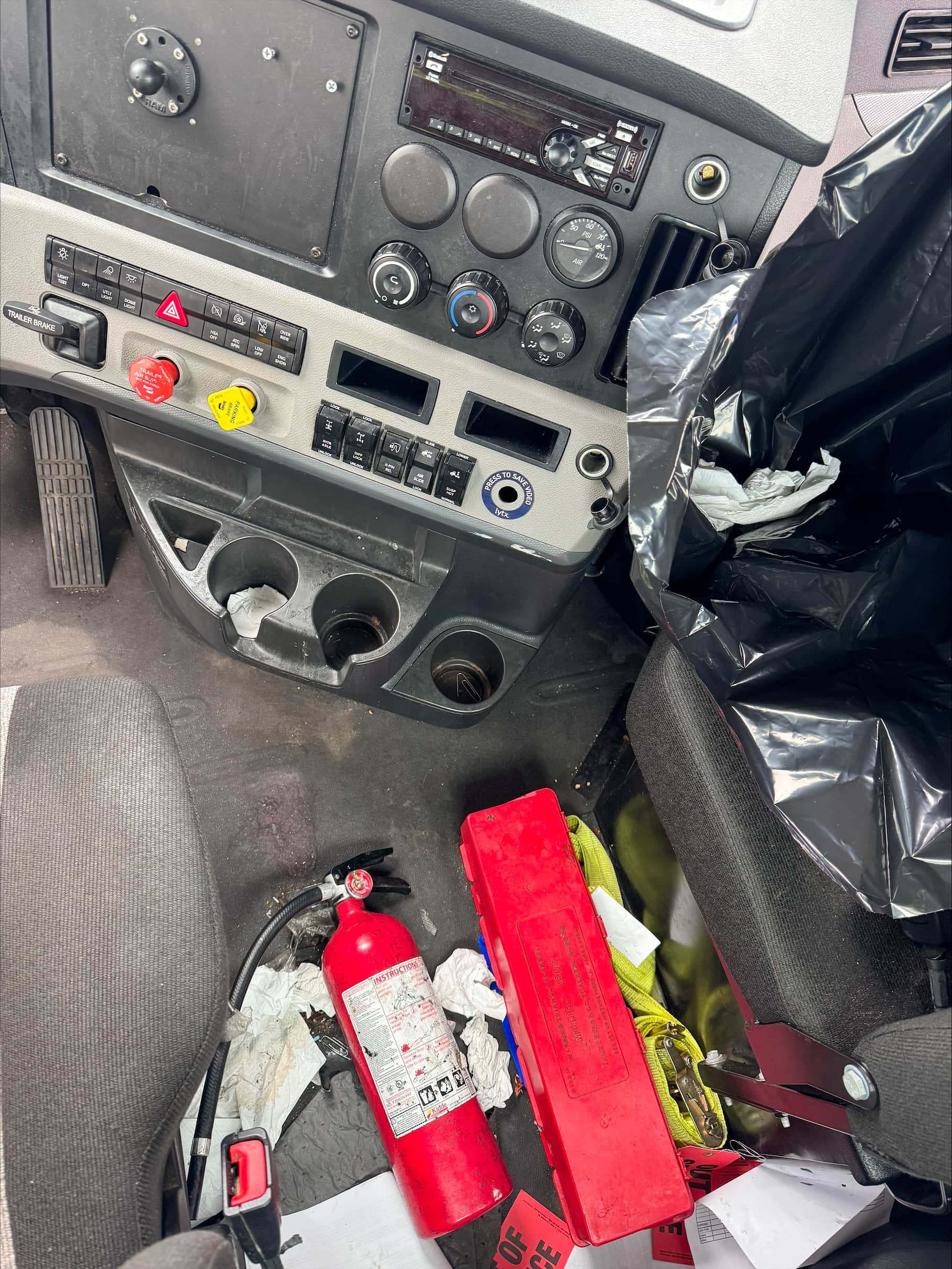 Interior of a vehicle showing the dashboard with control buttons and knobs, cup holders, and the floor cluttered with a red fire extinguisher, a red toolbox, papers, and a trash bag full of garbage.