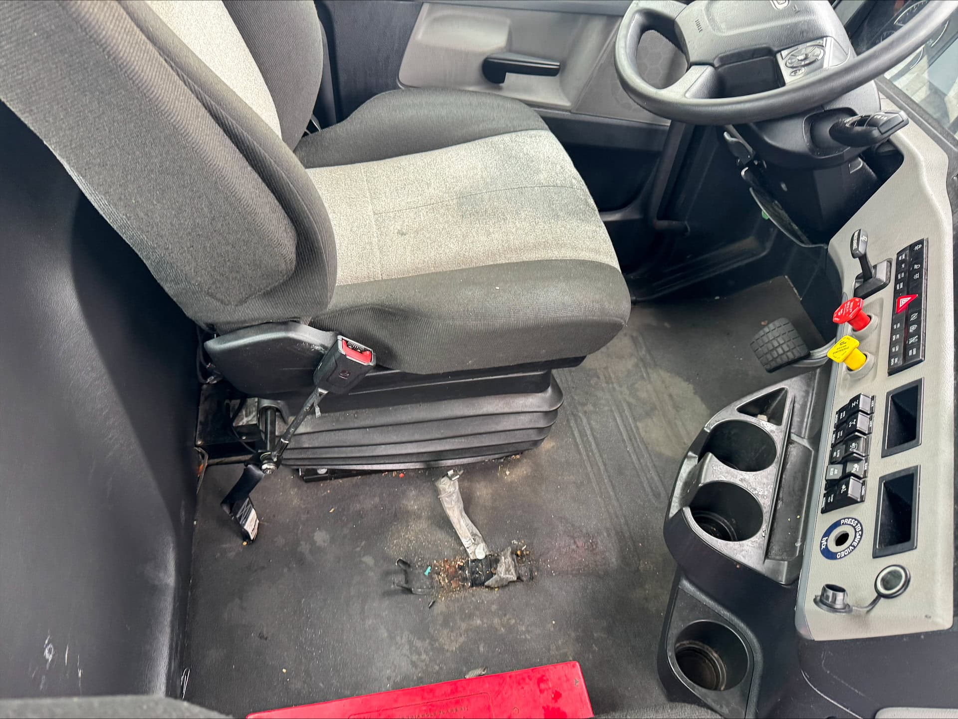 Drivers seat and control panel inside a vehicle, with worn upholstery, dirty floor, cup holders, switches, a yellow lever, and a red mat partially visible at the bottom of the image.