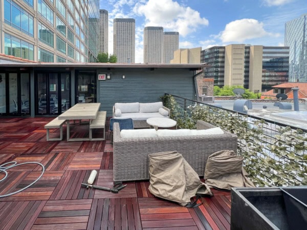A rooftop patio with wooden flooring, outdoor sofas, a dining table, and some chairs covered with beige covers. Modern office buildings are visible in the background under a partly cloudy sky.