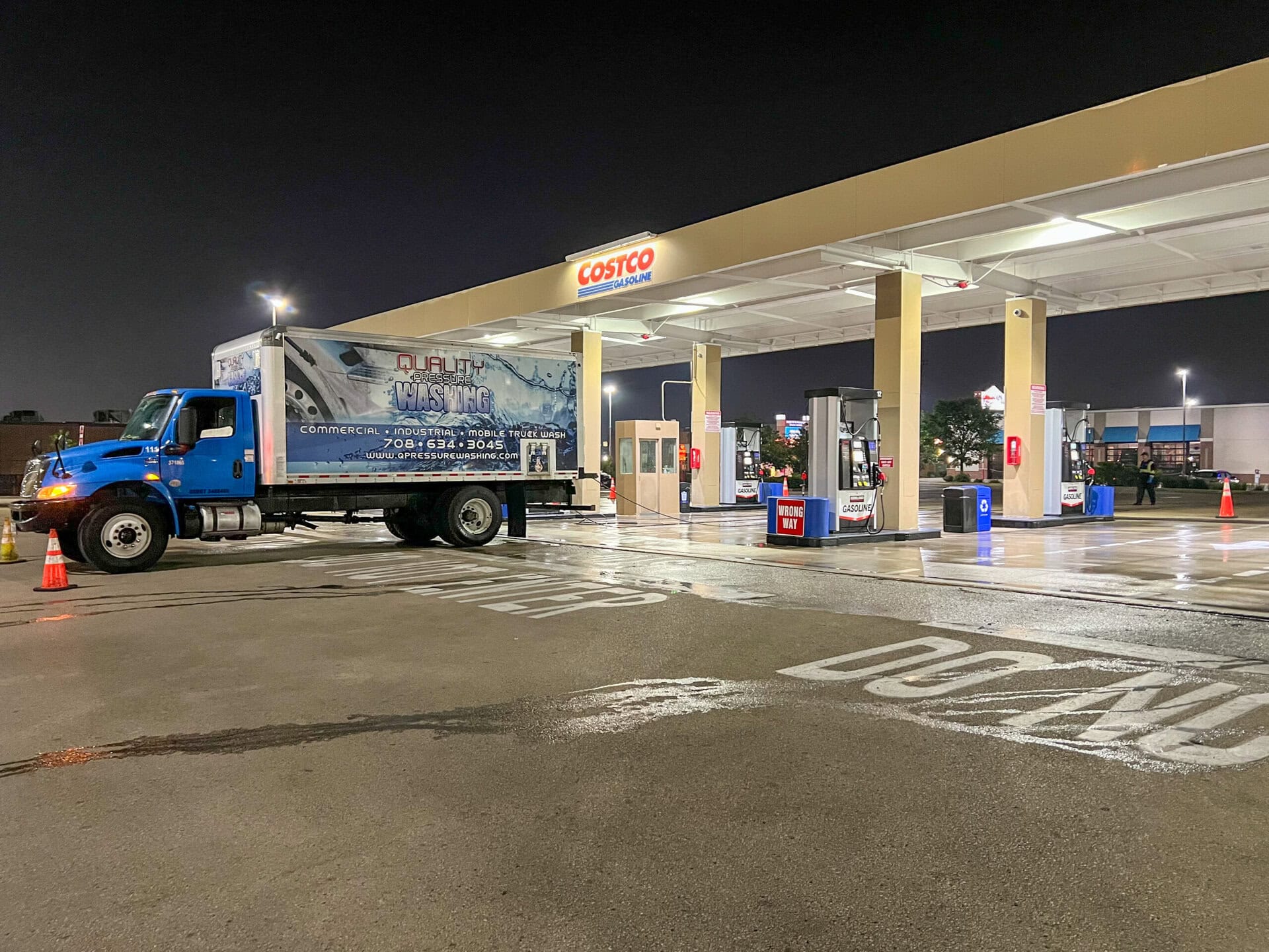 A blue Quality Power Washing truck is parked at a Costco gas station at night. Traffic cones are set up around the truck, and the area appears wet from cleaning. The station is well-lit and mostly empty.