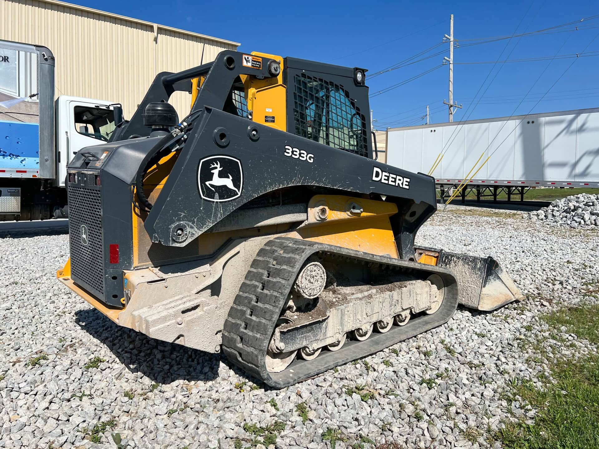 A John Deere 333G compact track loader sits on gravel near industrial buildings and trailers, with its bucket lowered and sunlight casting clear shadows.