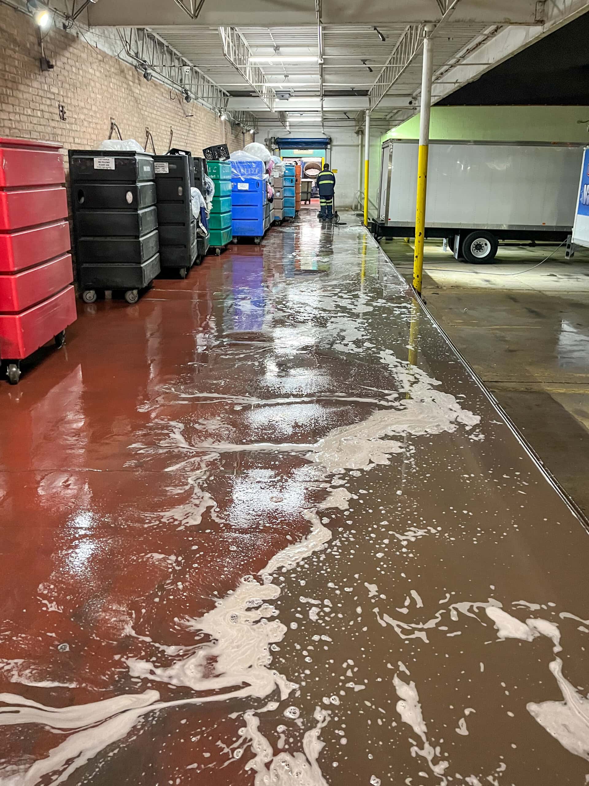 A corridor with a wet, soapy floor, lined with large carts and bins. A person in a safety vest is visible in the distance near a trailer, and overhead lights reflect off the shiny, reddish floor.
