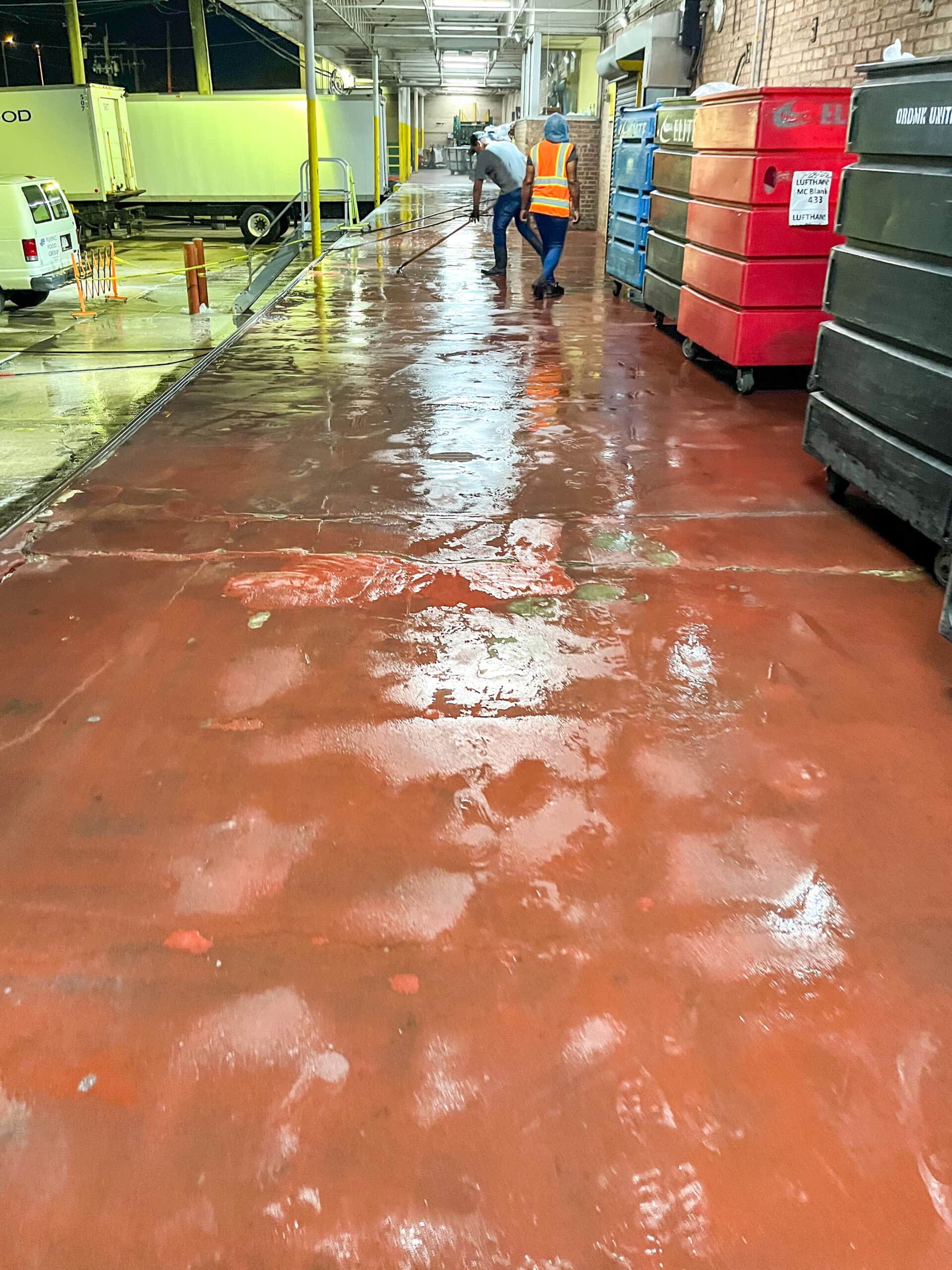 Two workers in safety vests clean a wet, red concrete loading dock next to large bins and a wall at night. The surface reflects lights, and a truck is visible in the background.