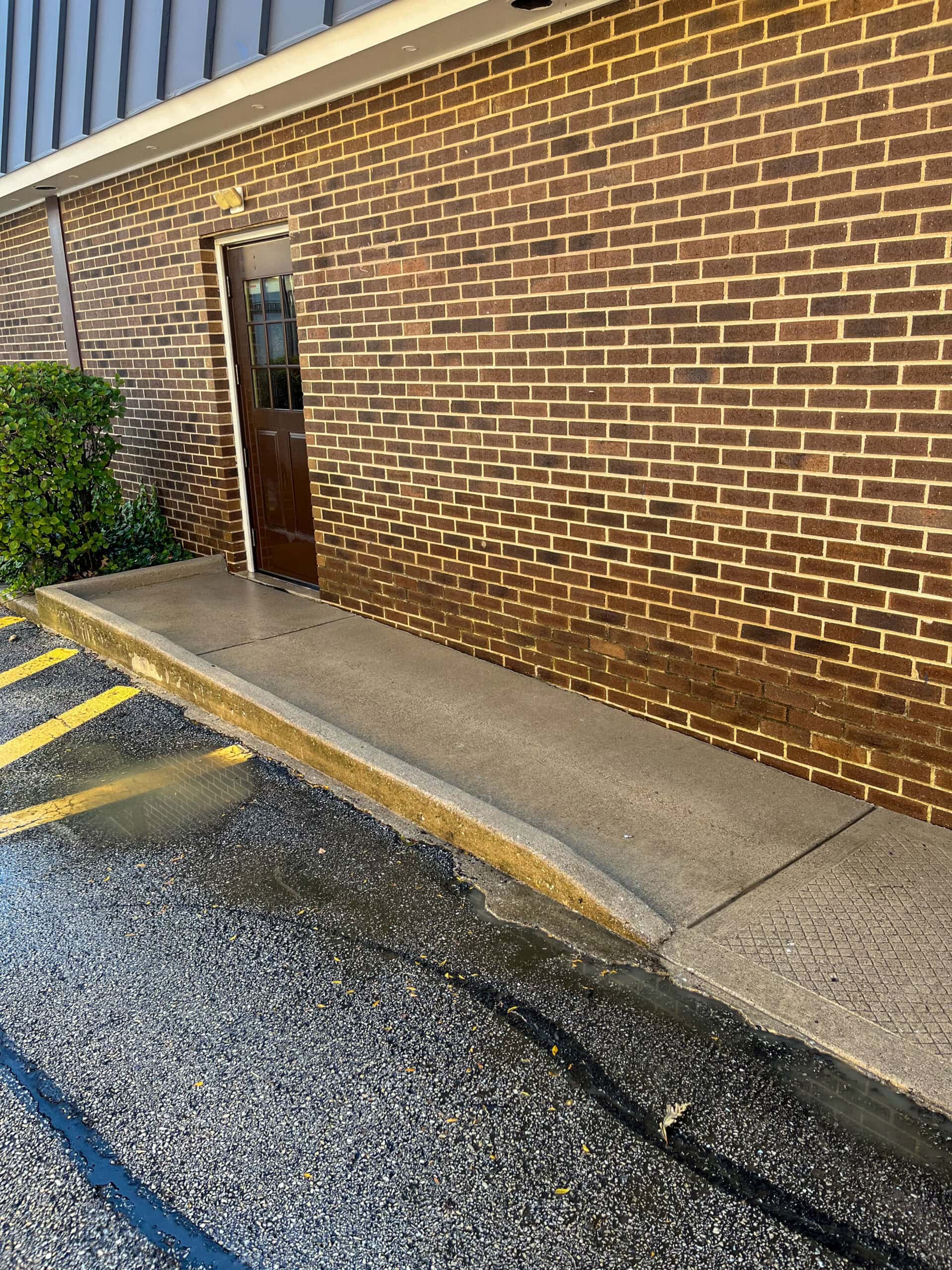 A brown brick building with a single closed door, a narrow concrete sidewalk in front, and yellow diagonal lines painted on the adjacent wet asphalt. A small green bush is on the left side near the wall.