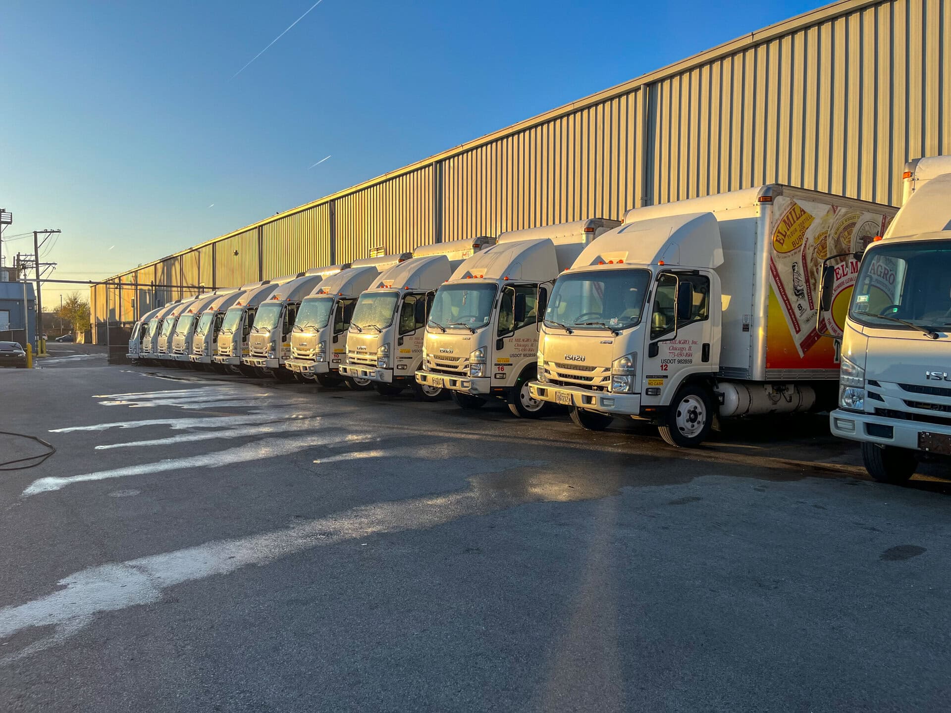 A row of white delivery trucks is parked side by side along a large industrial building under clear blue skies at sunrise or sunset.