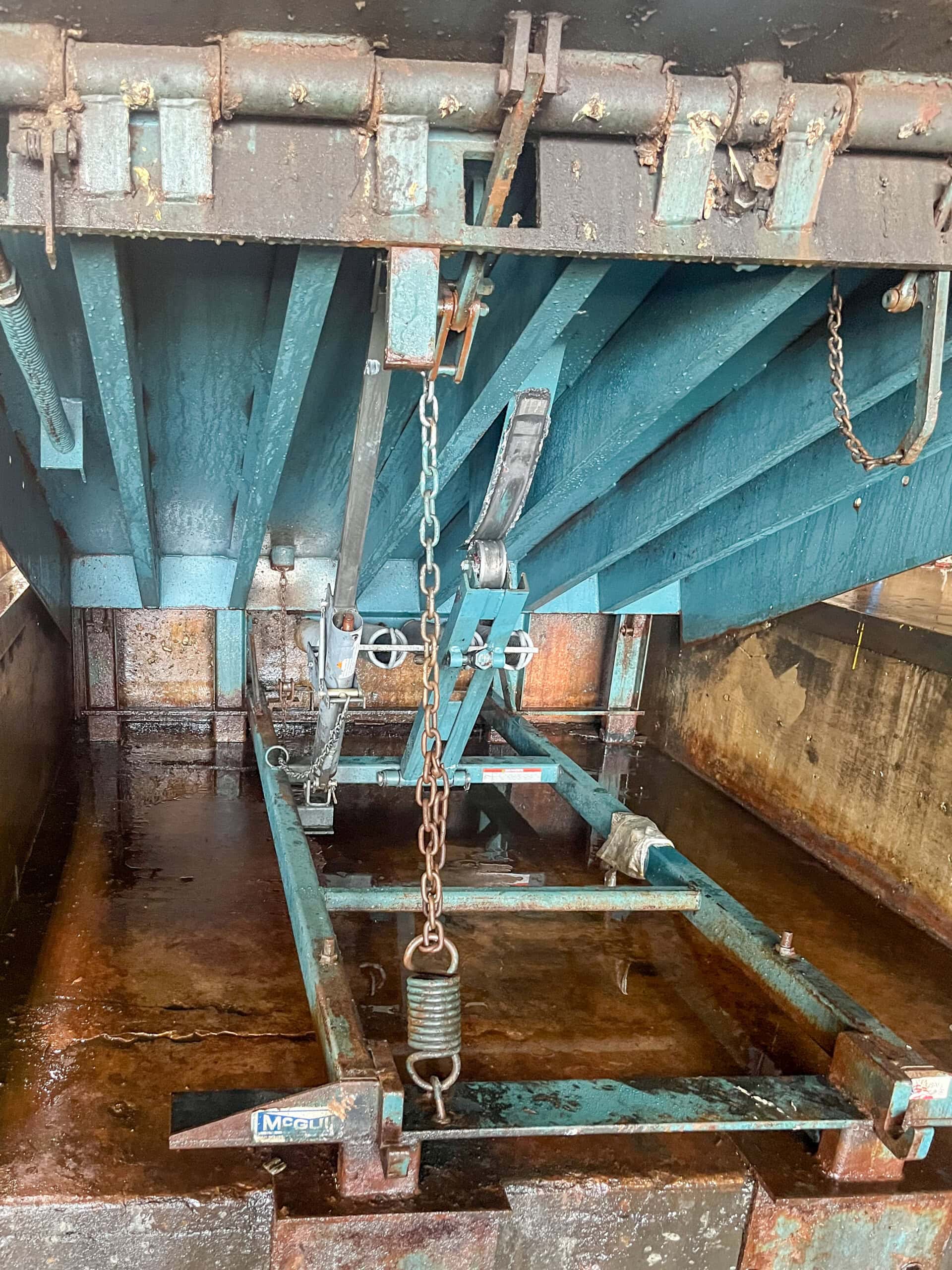 A close-up view of an industrial metal structure with teal beams, chains, springs, and levers, set against a rusted, metallic surface. The machinery appears to be part of a loading or lifting mechanism.
