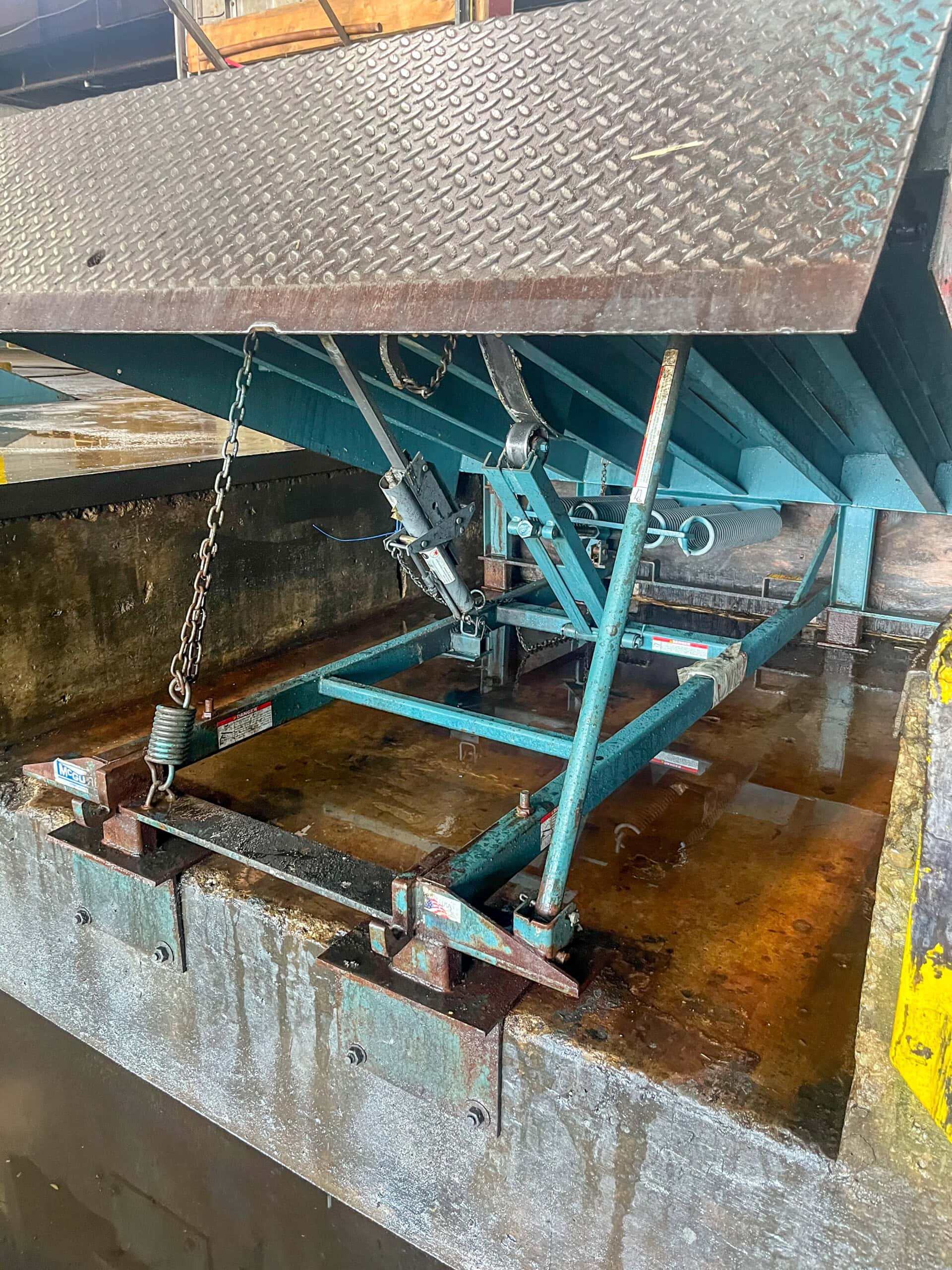 A close-up view of a loading dock leveler with a diamond plate steel ramp partially raised, showing metal support mechanisms, a spring, and a chain underneath. The area beneath appears wet and rust-stained.