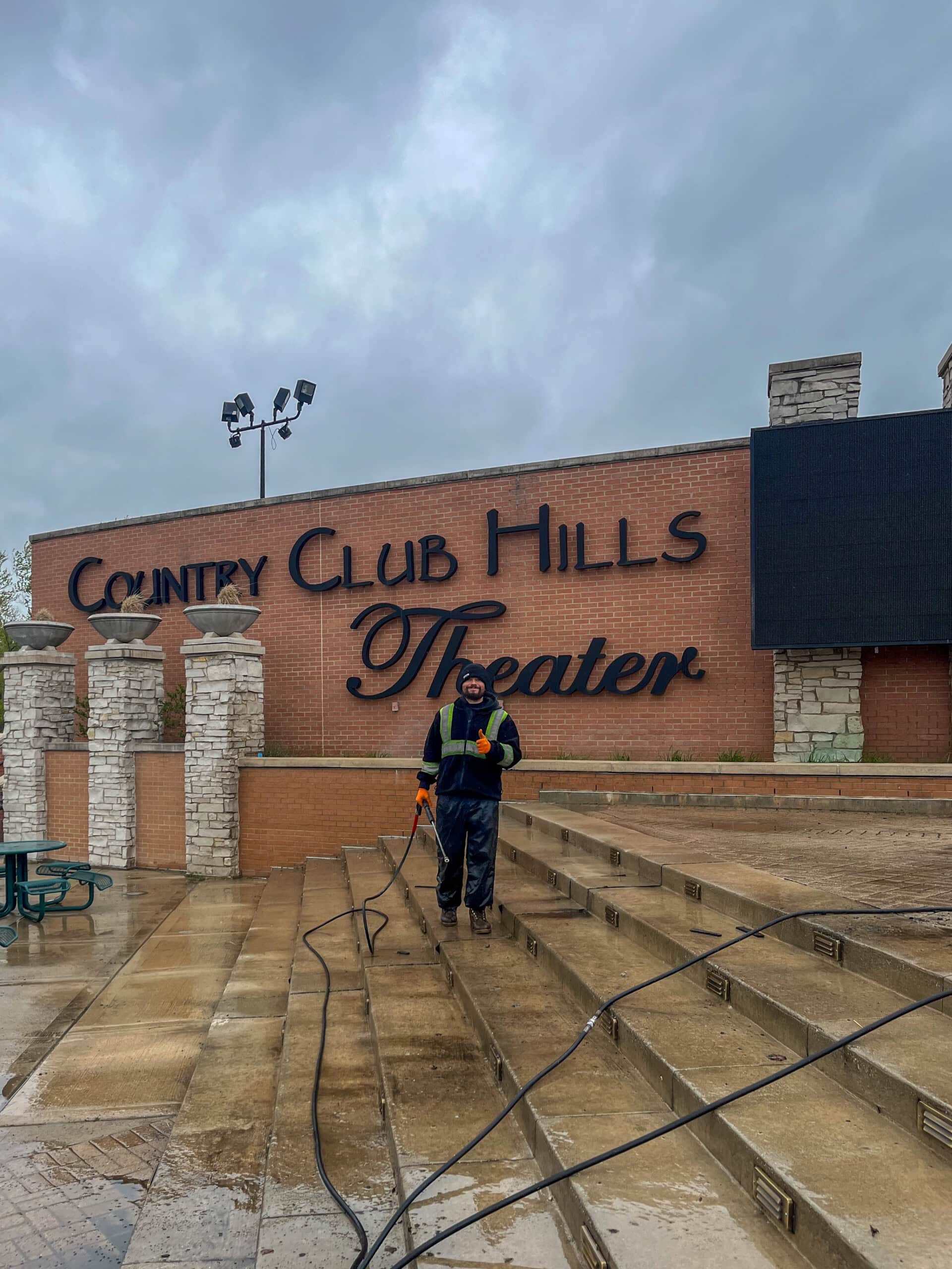 A person pressure washes outdoor steps in front of a brick building with a large Country Club Hills Theater sign on a cloudy day.