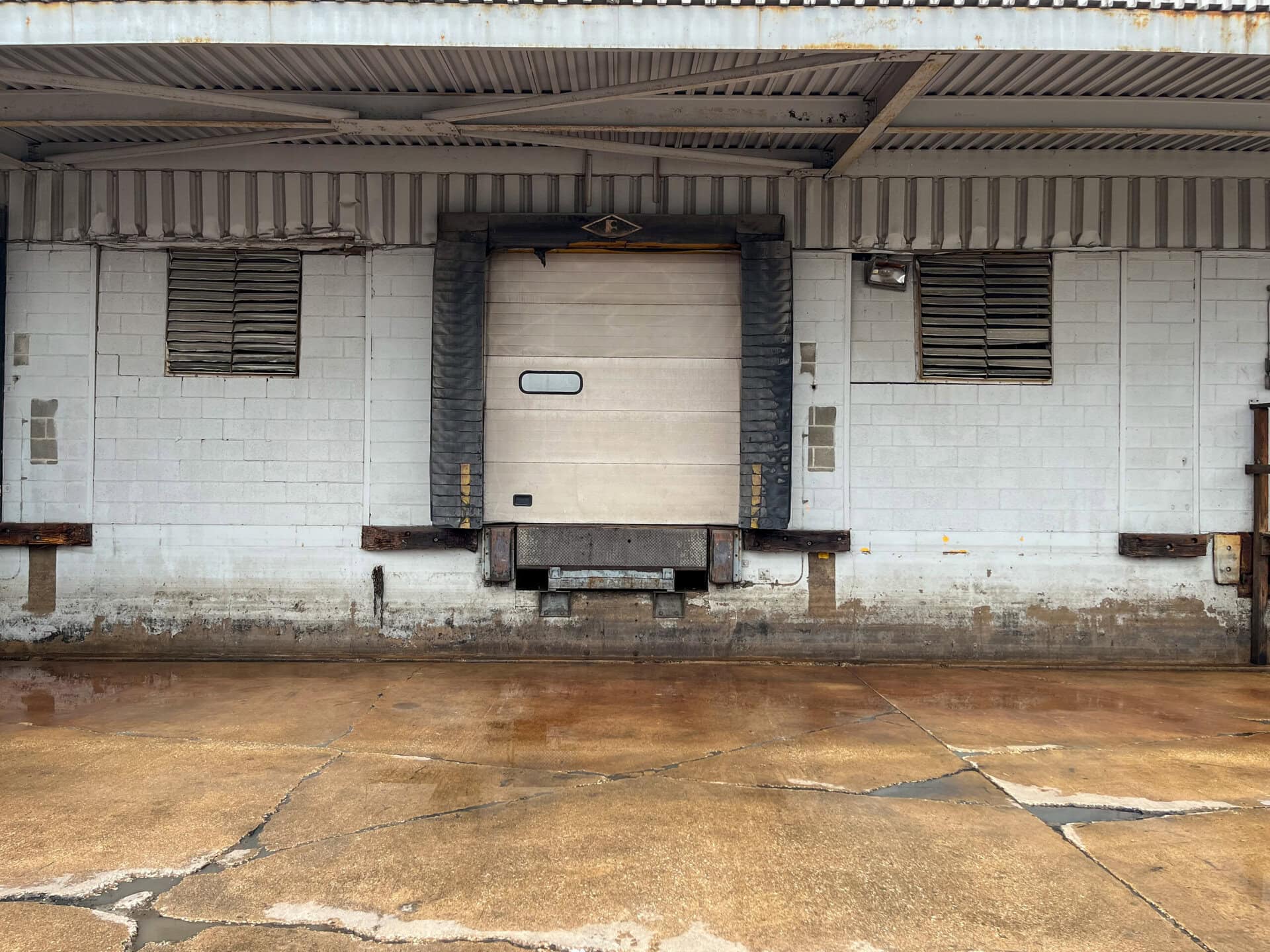 A closed loading dock door with black padding frames the opening. The wall is white with two barred windows and some weathered stains. The concrete ground in front shows cracks and wet patches.