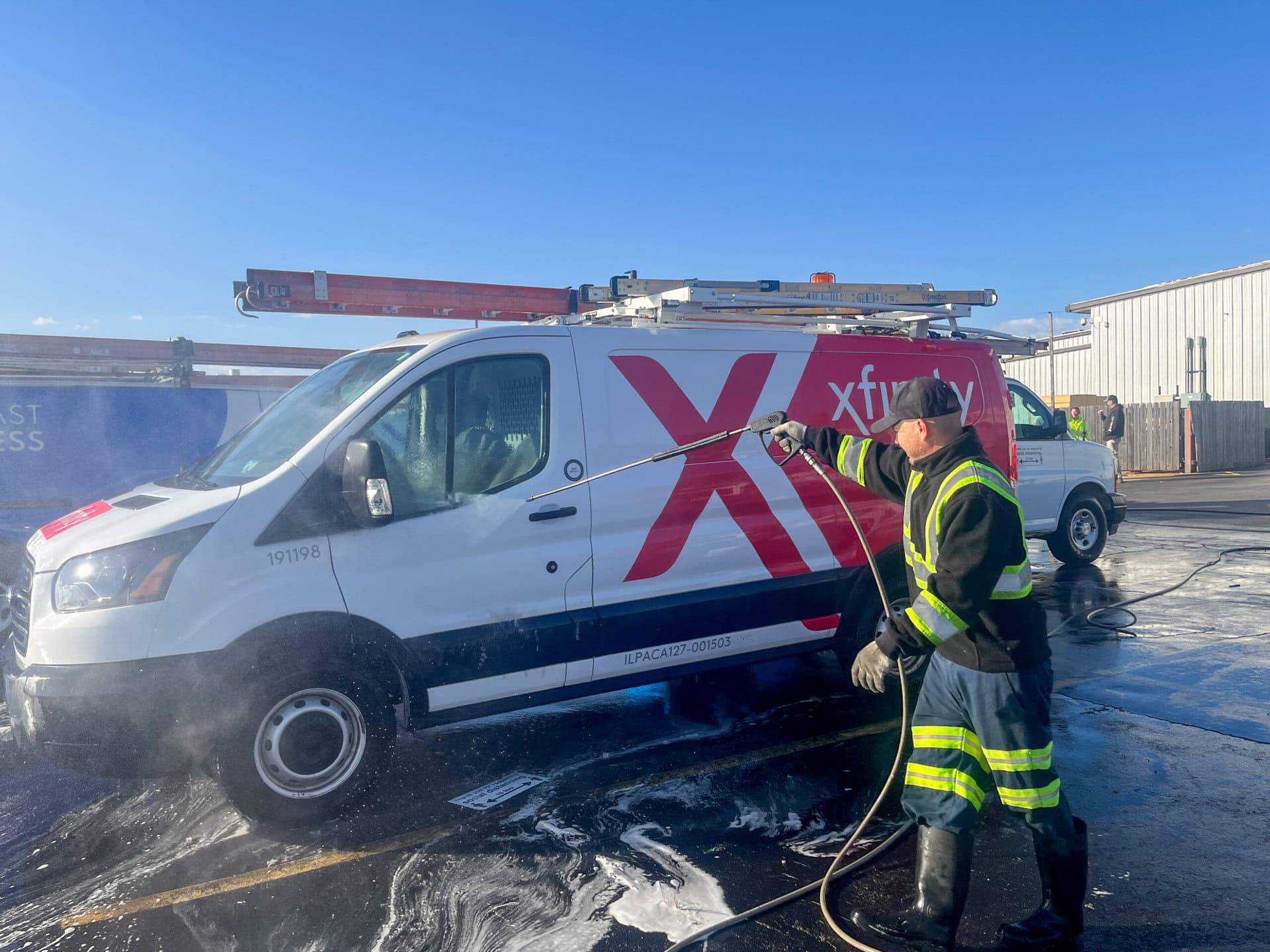 A worker in high-visibility clothing uses a pressure washer to clean an Xfinity service van in a parking lot on a sunny day.