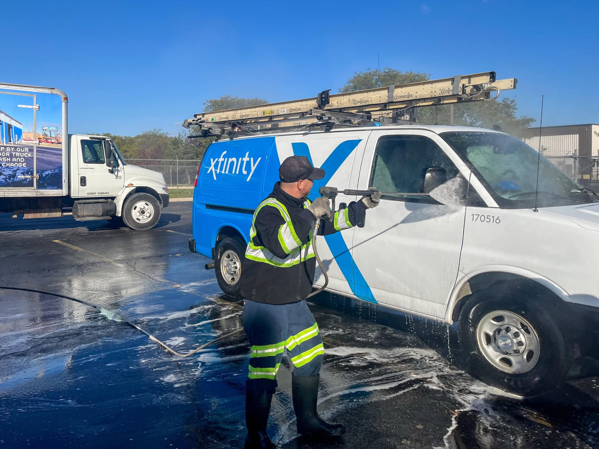 A worker in reflective gear uses a pressure washer to clean a white and blue Xfinity van in a parking lot on a sunny day. Soap and water are visible on the ground. Another truck is parked nearby.