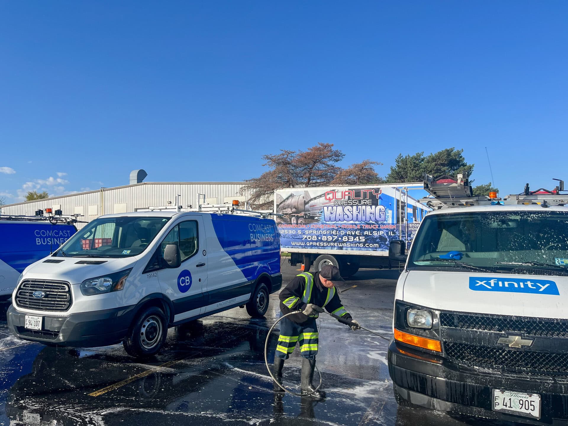A worker in safety gear uses a hose to wash a Comcast Business van and an Xfinity van in a parking lot, with a utility truck and a building in the background under a clear blue sky.