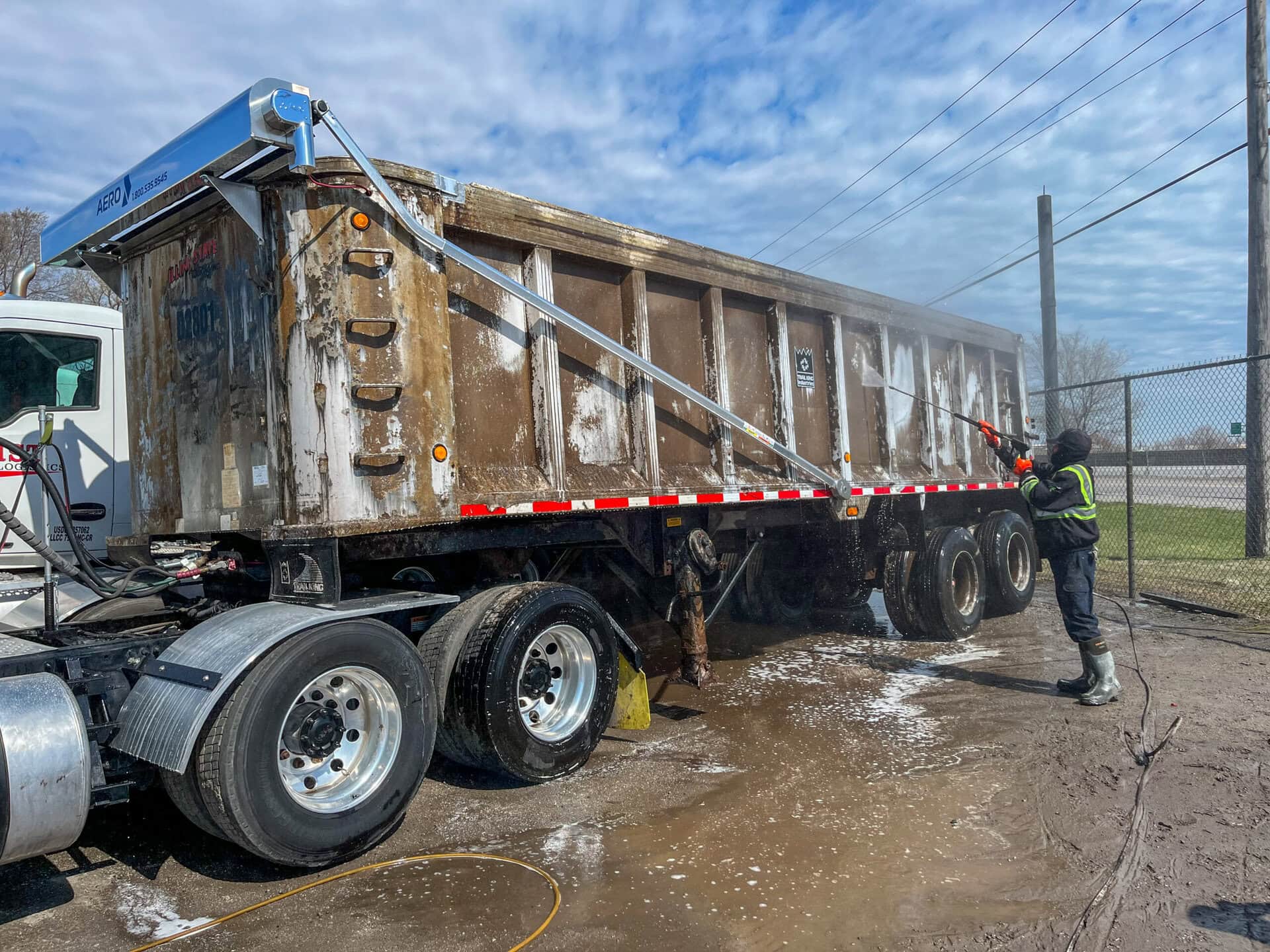 A worker in safety gear uses a power washer to clean a large, dirty dump truck trailer in an outdoor area on a cloudy day.