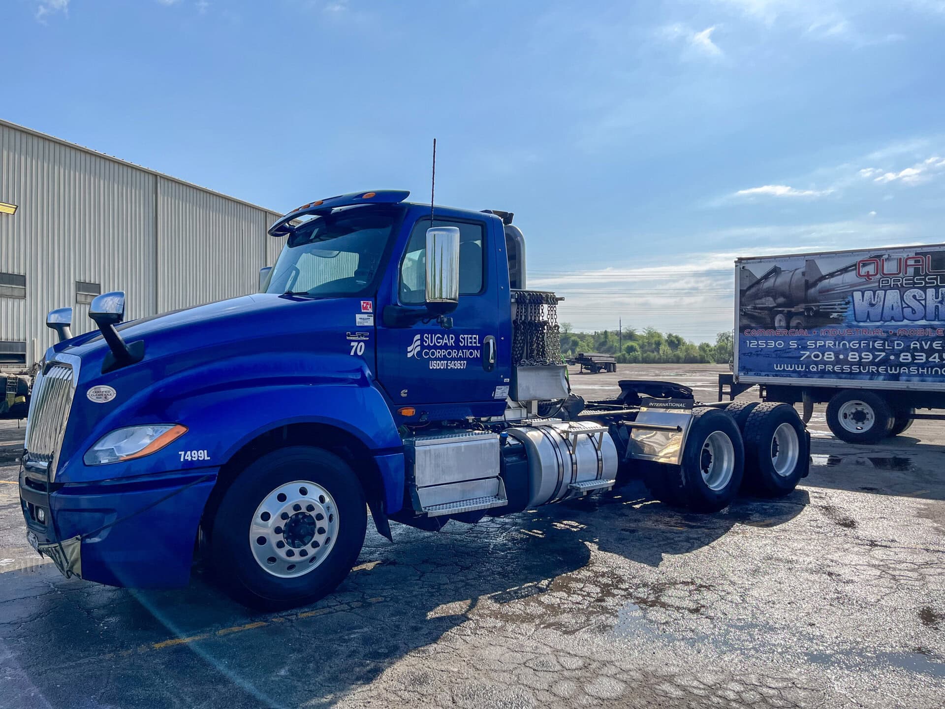 A blue semi-truck cab is parked on a lot near a warehouse. A box truck with a car wash advertisement is in the background. The sky is clear and the ground is wet, reflecting the vehicles.