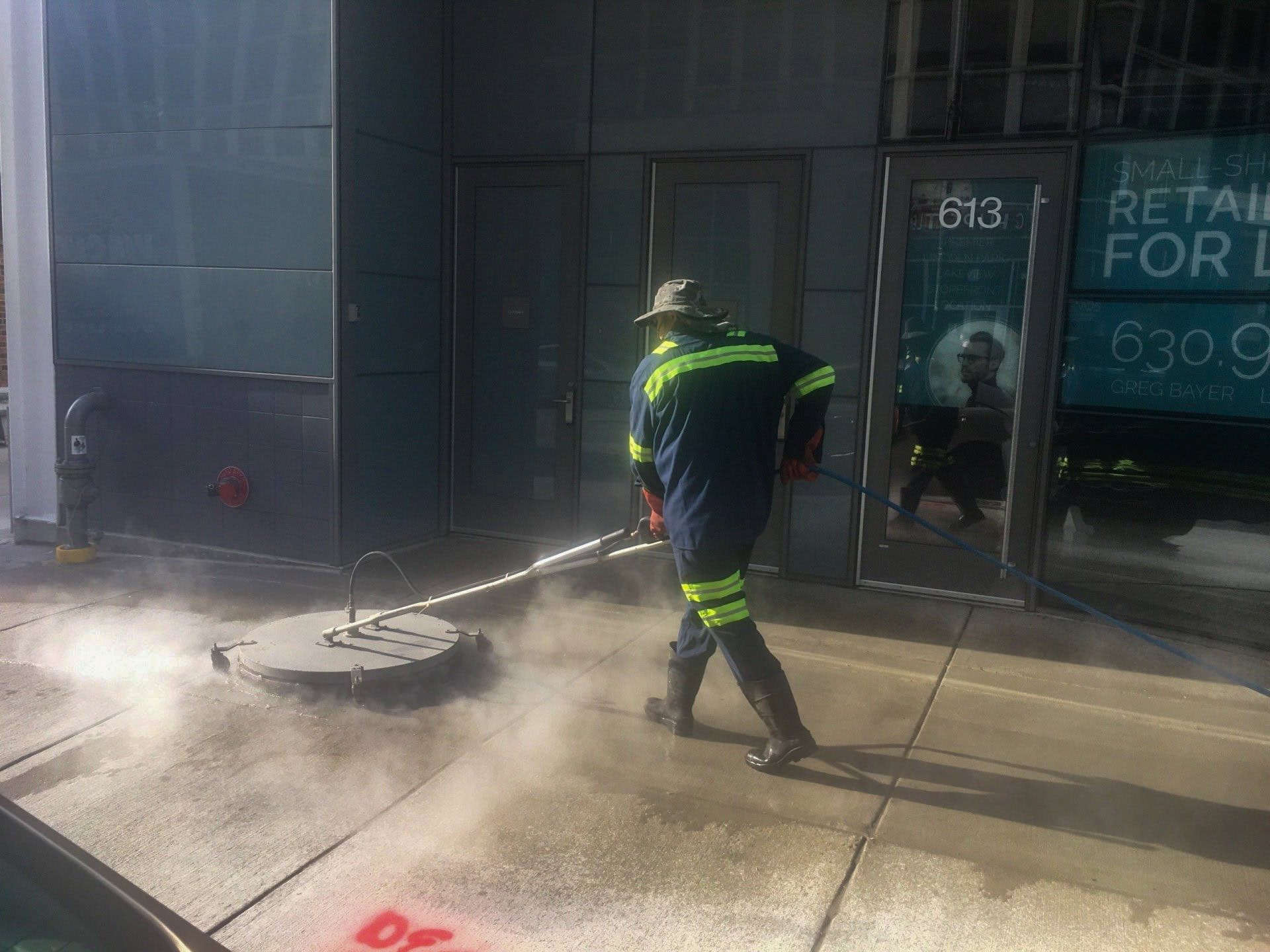 A person wearing protective gear uses a large power washer to clean a city sidewalk, with steam rising from the pavement. Glass doors and retail signs are visible in the background.
