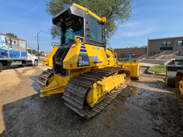 A bright yellow Komatsu bulldozer with mud on its tracks stands on a construction site near a building and a truck, with a tree and blue sky in the background.