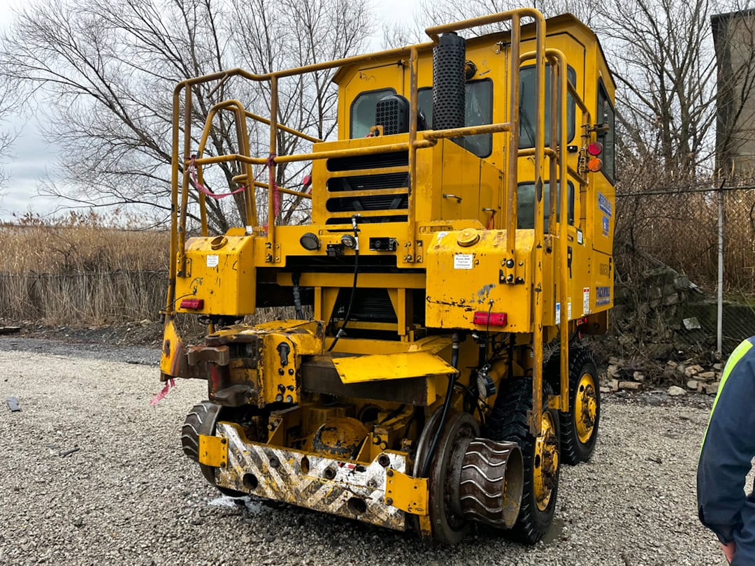 A large yellow maintenance vehicle with rail wheels and heavy machinery parts is parked on gravel near a fence and leafless trees, under a cloudy sky.