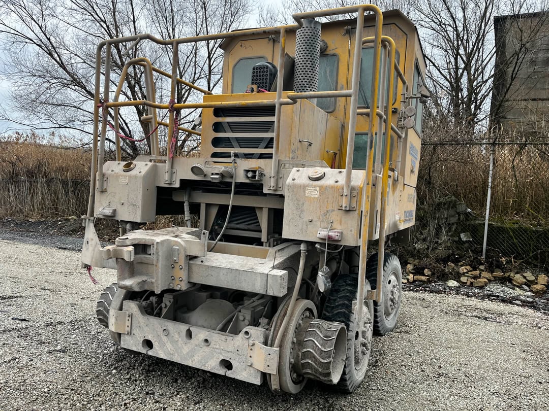 A large, industrial yellow rail maintenance machine with metal railings, multiple wheels, and heavy-duty components sits on a gravel lot, surrounded by dry grass, leafless trees, and a chain-link fence.