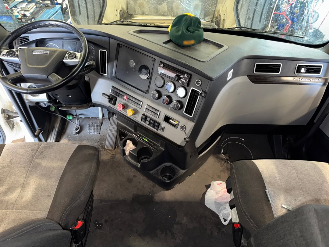 Interior cab of a Freightliner Cascadia truck, showing the steering wheel, dashboard with various controls and buttons, two seats, cup holders, and a green hat placed on the dashboard. A plastic bag is on the floor.