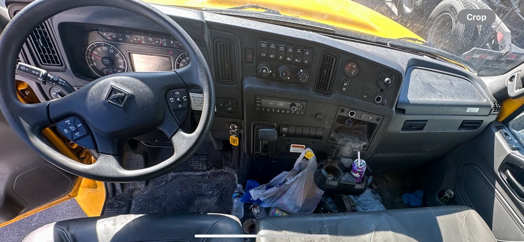 The interior of a yellow truck’s cab, showing the steering wheel, dashboard with various controls, pedals, and a cluttered center area with trash, a cup, and a plastic bag on the floor.
