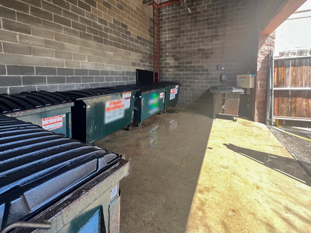 Four large dumpsters with black lids are lined up against a brick wall in a concrete-walled outdoor area. Sunlight partially brightens the scene, and a wooden gate is open on the right side.