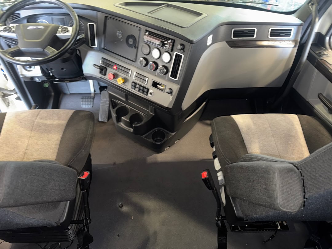 The interior cab of a truck, showing two gray seats, a steering wheel on the left, multiple control buttons and switches on the dashboard, and cup holders between the seats.