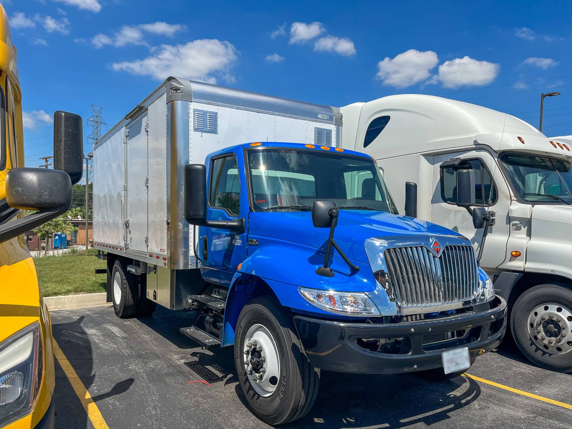 A bright blue box truck is parked in a lot between a yellow vehicle and a white semi-truck under a sunny, blue sky with scattered clouds.