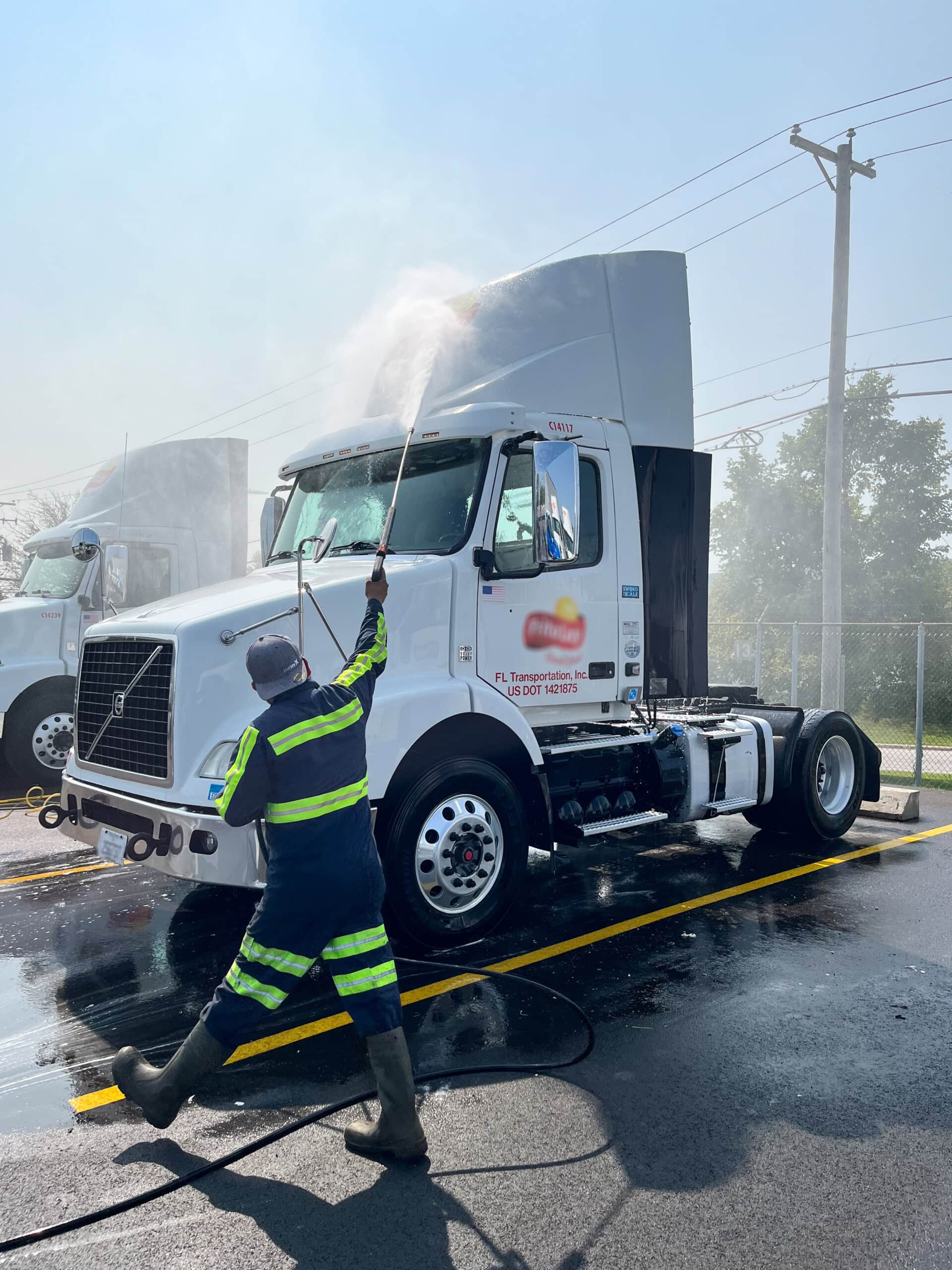 A worker in a reflective uniform and boots uses a high-pressure hose to wash the cab of a white semi-truck parked on wet pavement, with another truck and trees visible in the background.