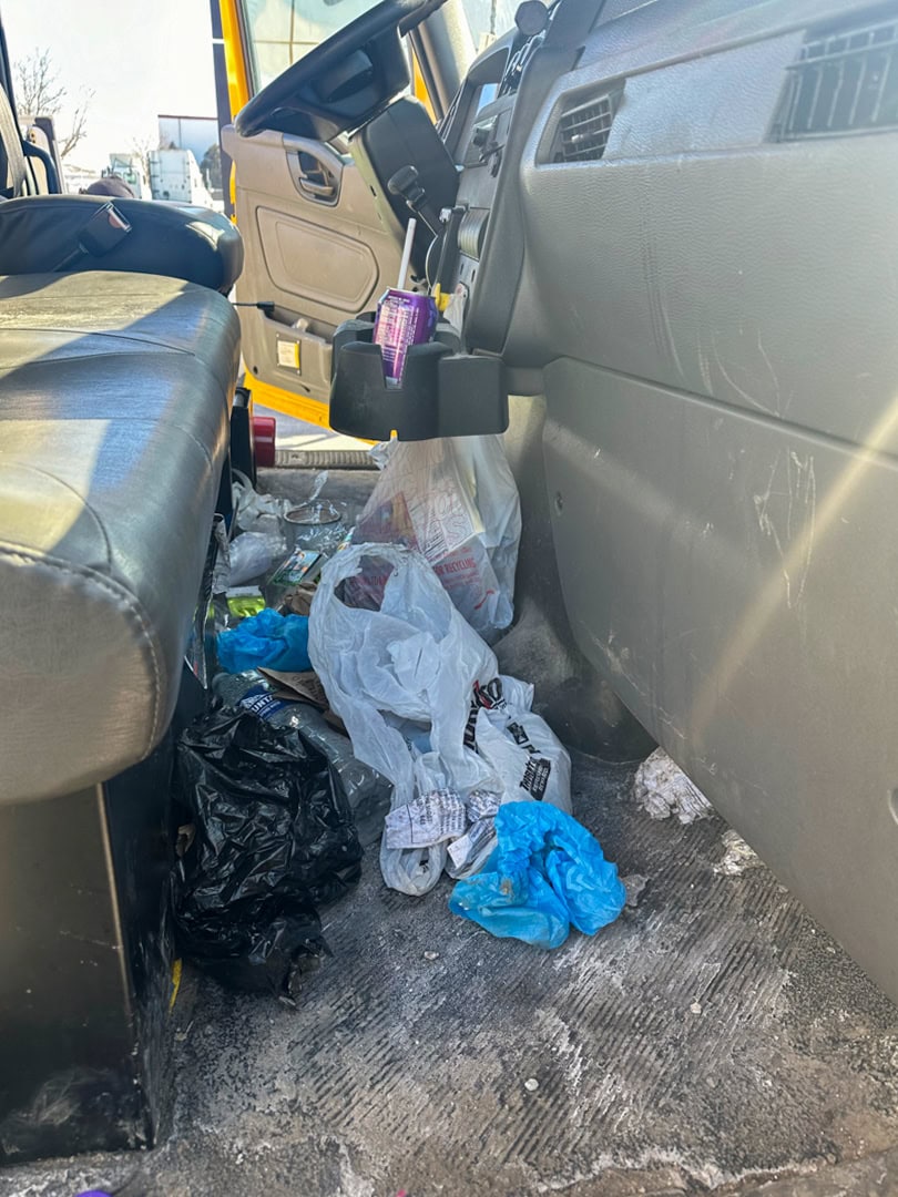 Trash, plastic bags, gloves, and cans litter the floor of a vehicles front passenger area, while sunlight shines through the open door. The vehicle interior looks cluttered and unclean.
