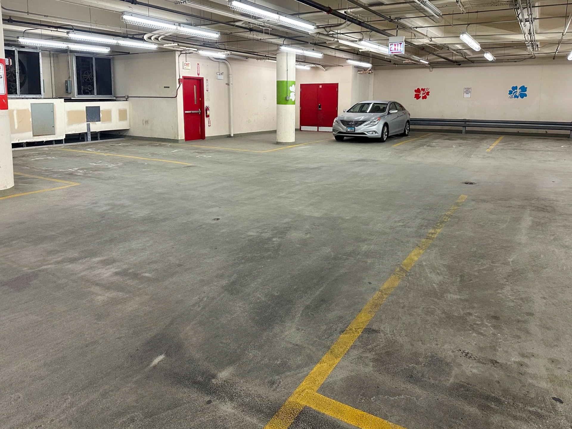A mostly empty indoor parking garage with one silver car parked near the back wall. The space is brightly lit with overhead lights, and there are red doors and colored wall markings visible.