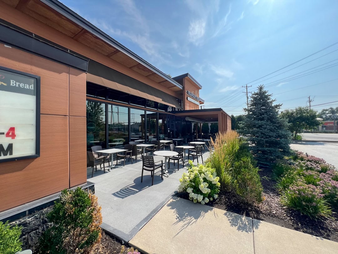 Outdoor seating area of a modern café or restaurant with empty tables and chairs, surrounded by landscaped plants and flowers, on a sunny day with a clear blue sky.