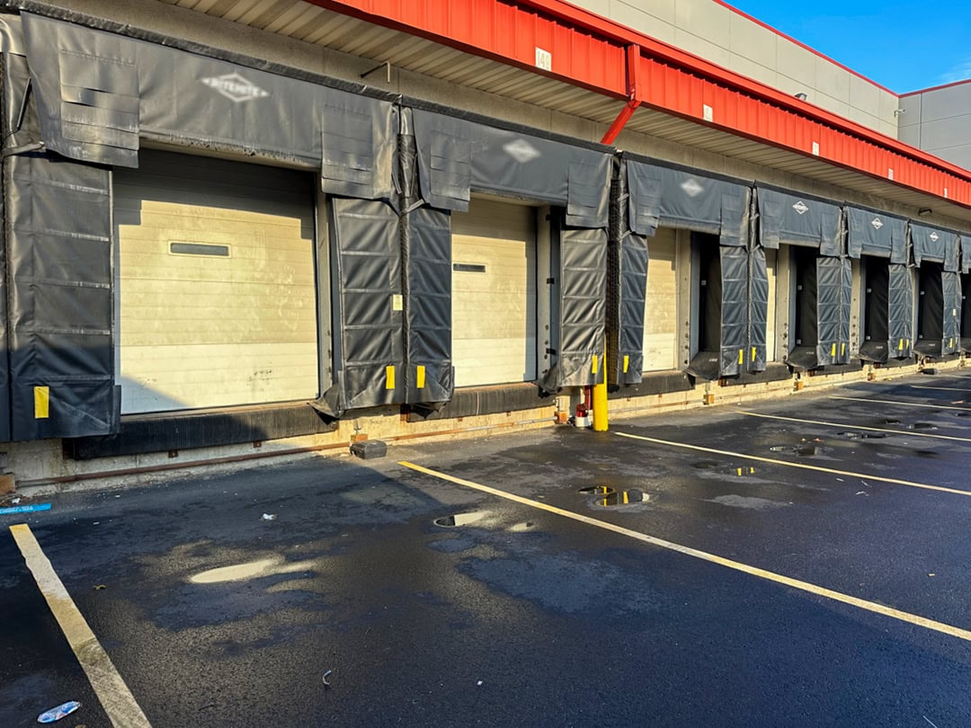 A row of closed loading dock doors at a distribution center, with black protective padding and yellow bumpers, stands beside an empty, wet asphalt parking area under a blue sky.