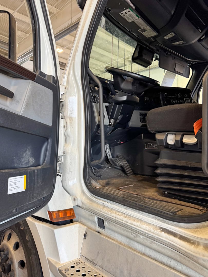 Open driver-side door of a white semi-truck shows the interior cabin, driver’s seat, foot pedals, steering wheel, and dashboard; exterior is slightly dirty and the truck is parked indoors.