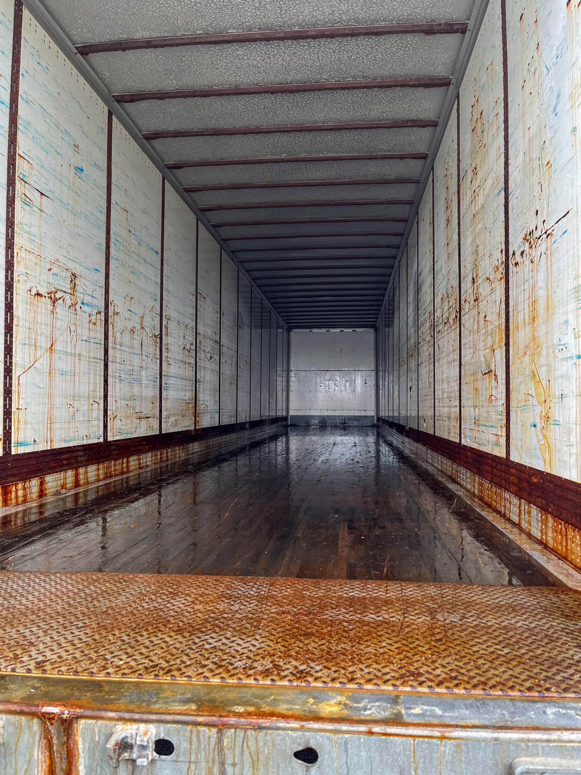 The image shows the inside of an empty, dirty freight trailer with rusty streaks on the walls and floor, and standing water scattered across the wooden flooring. The trailer doors are open, revealing its full length.