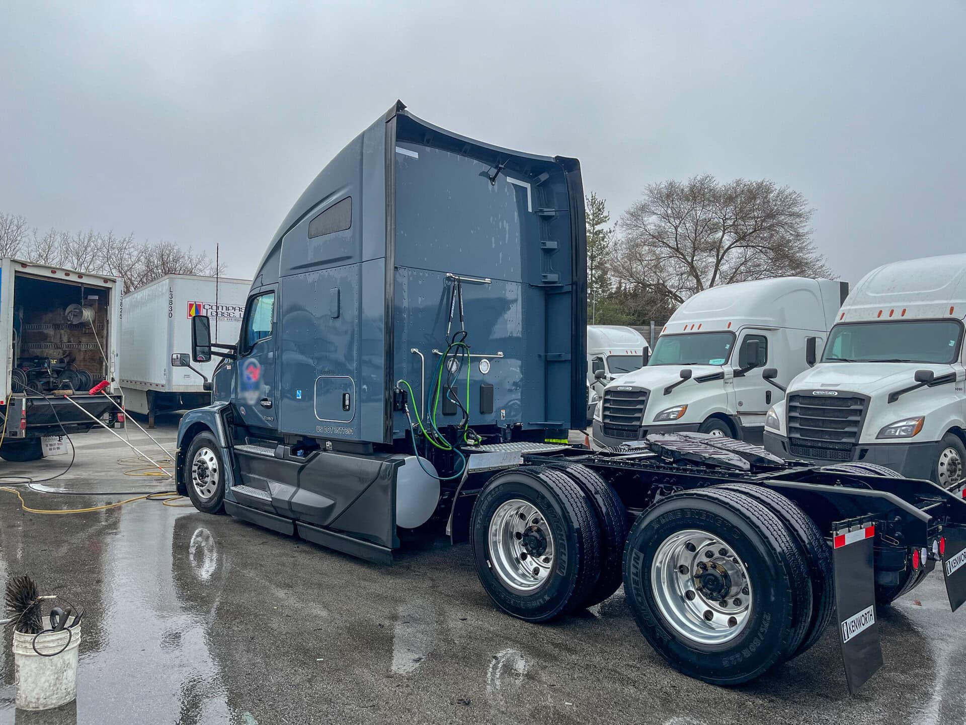 A dark blue semi-truck cab parked on wet pavement near other white trucks, with hoses attached and trees in the background under an overcast sky.