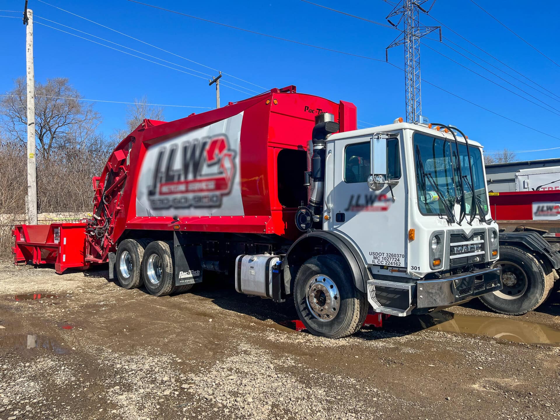 A large red and white garbage truck is parked on a gravel lot under a clear blue sky. The truck has a company logo on the side and another garbage truck is visible in the background.