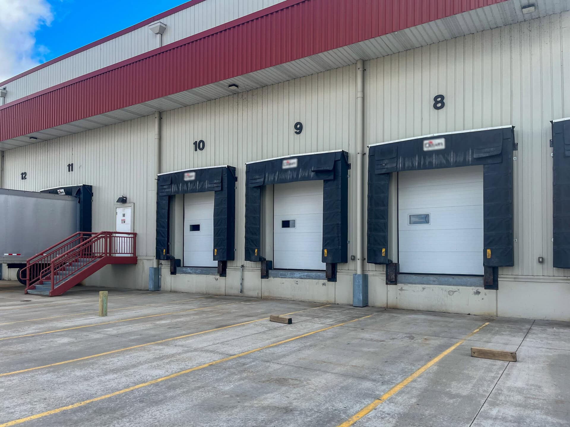 Four loading dock doors numbered 8, 9, 10, and 11 at a warehouse, with a trailer parked at door 11 and red stairs to the left. The ground has yellow parking lines and concrete wheel stops.