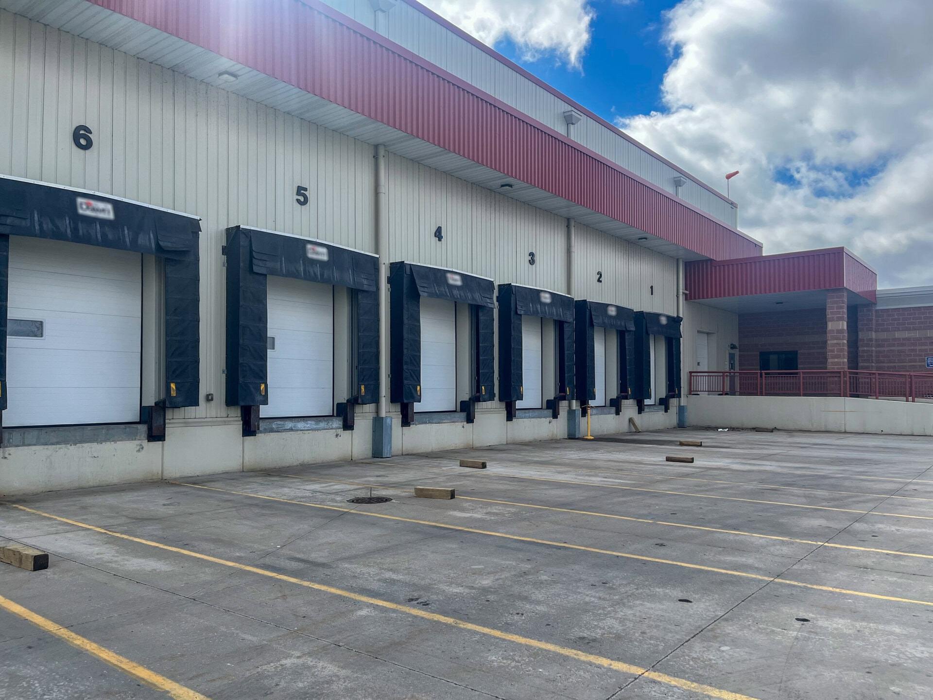 A row of six closed loading dock doors, each numbered, at a warehouse or distribution center with an empty, marked parking area under a partly cloudy sky.