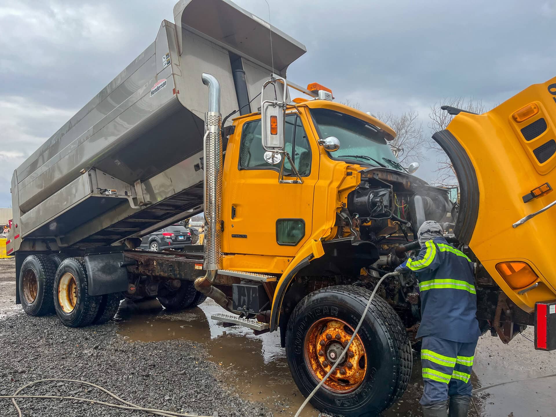 A worker in a reflective suit sprays water at the exposed engine of a large yellow dump truck with its hood open and bed raised, in an outdoor, muddy area under cloudy skies.