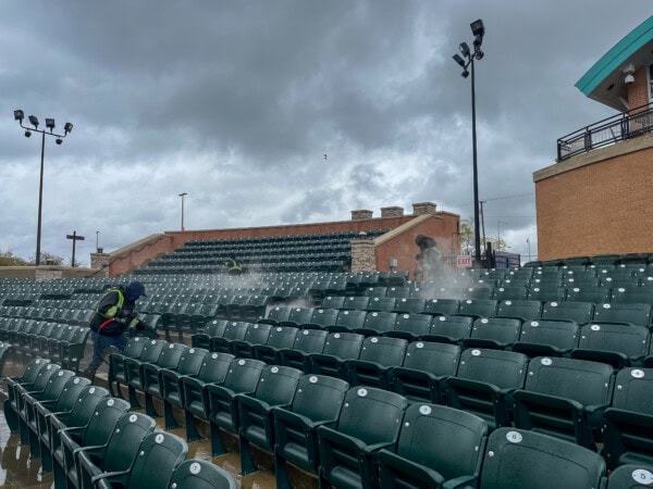Two workers in safety vests use power washers to clean rows of empty green stadium seats under a cloudy, overcast sky. Water mist is visible rising from the seats. The stadium is empty and wet.