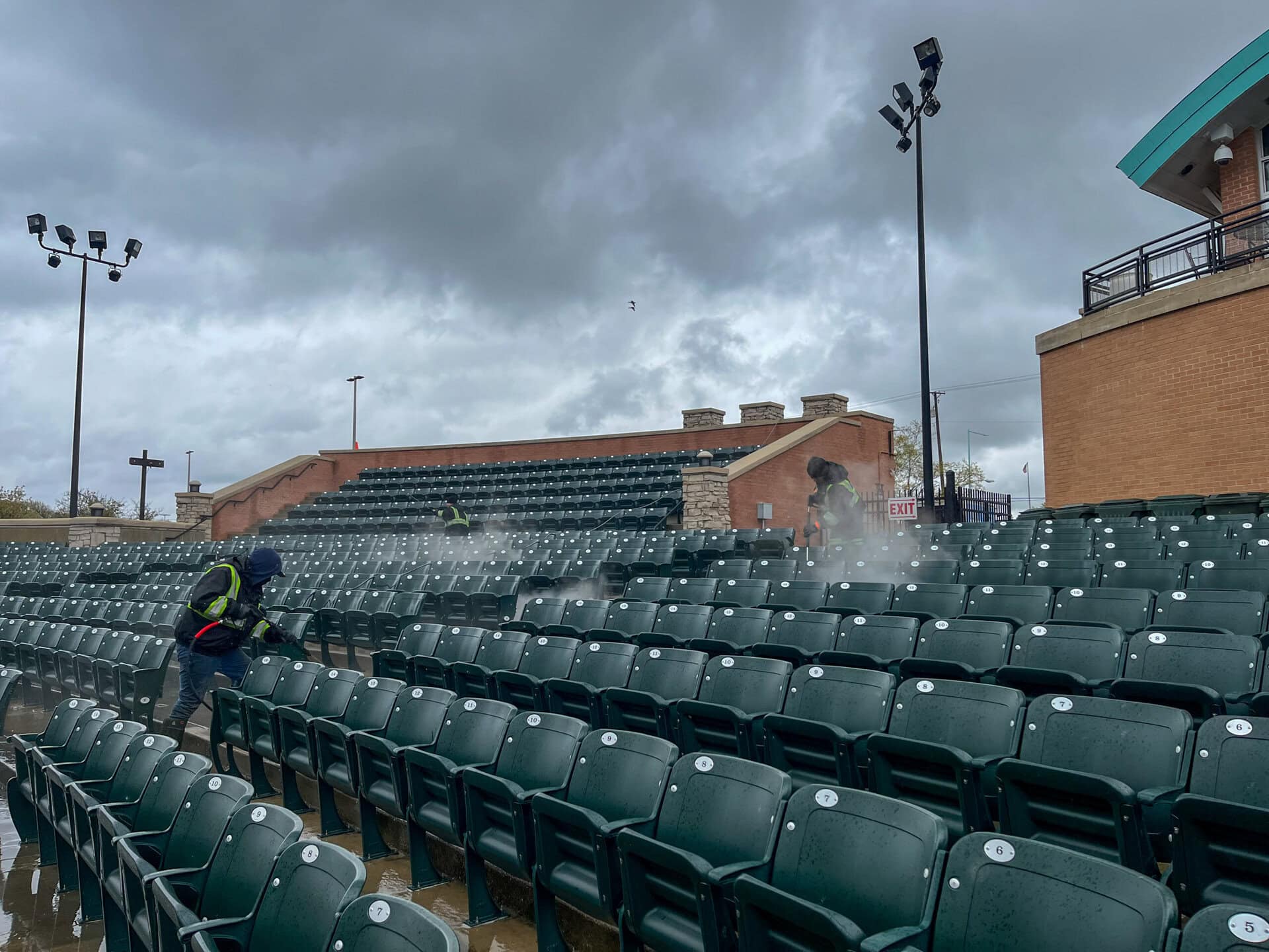 Two workers in safety vests use power washers to clean rows of empty green stadium seats under a cloudy, overcast sky. Water mist is visible rising from the seats. The stadium is empty and wet.
