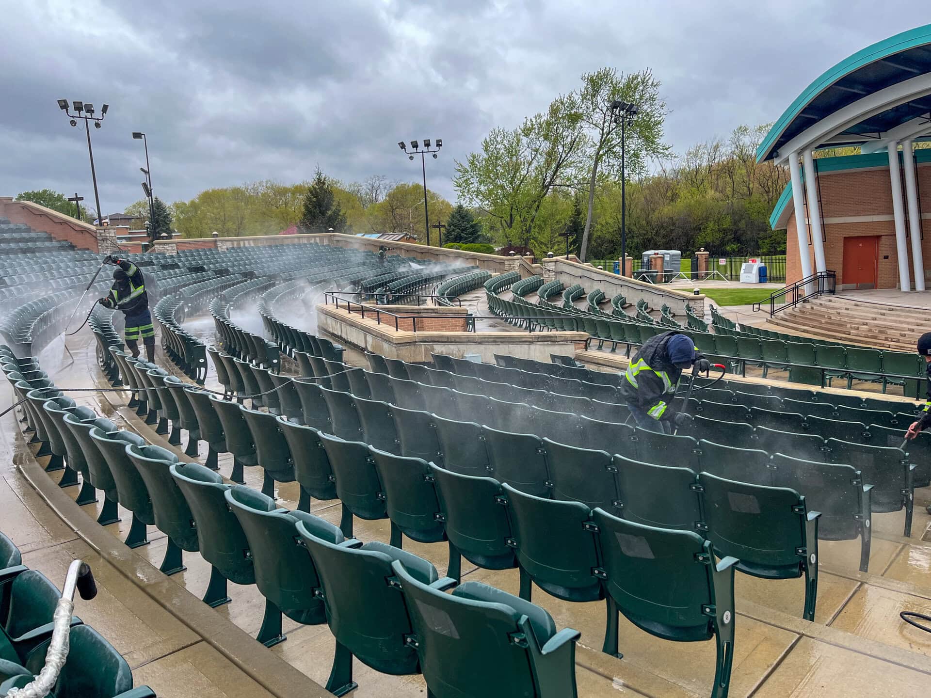 Workers in safety gear use pressure washers to clean rows of green seats in an outdoor amphitheater. The area is wet, and trees and a covered stage are visible in the background under a cloudy sky.