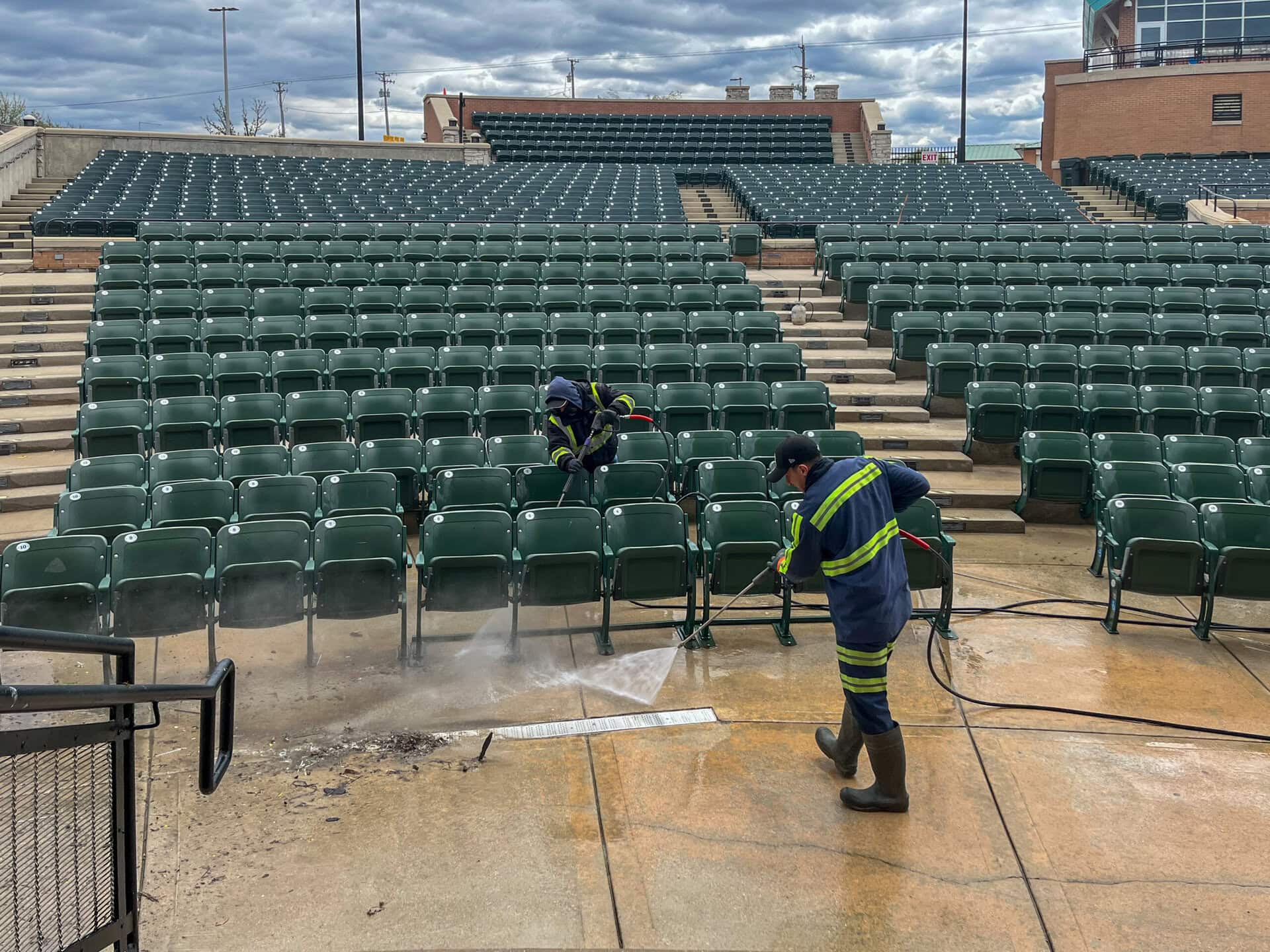 Two workers in safety gear use power washers to clean the concrete floor in front of empty green stadium seats on a cloudy day.