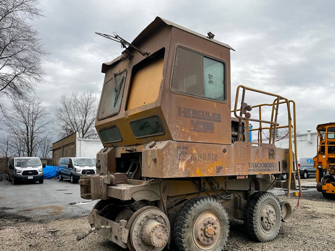 A weathered, rusted railcar mover vehicle with Hercules and Trackmobile branding sits on gravel near parked vans under a cloudy sky. Leafless trees and a building are in the background.