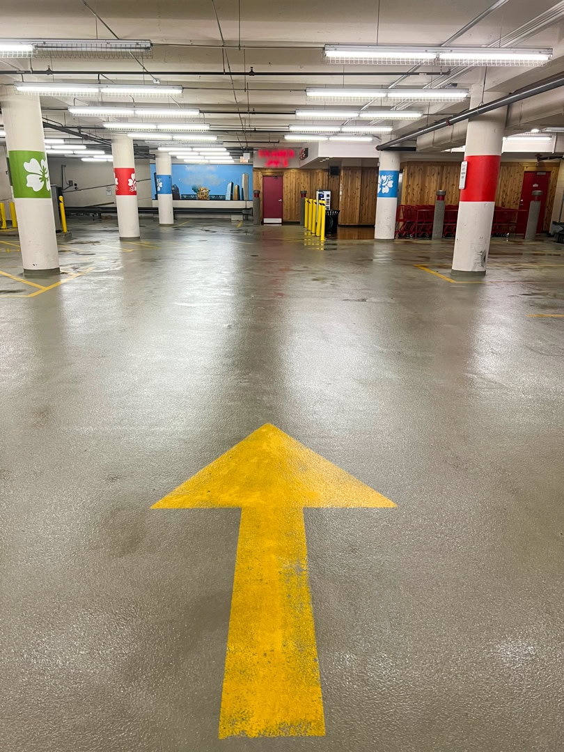 A yellow arrow is painted on the floor of a clean, brightly lit underground parking garage with red and white pillars and signs on the walls.