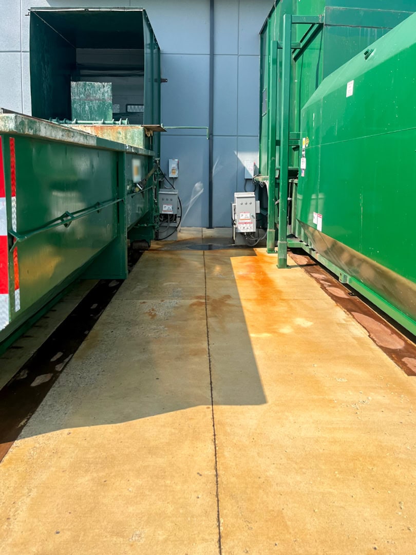A concrete pathway with rust stains runs between two large green industrial dumpsters beside a gray building wall under bright sunlight.