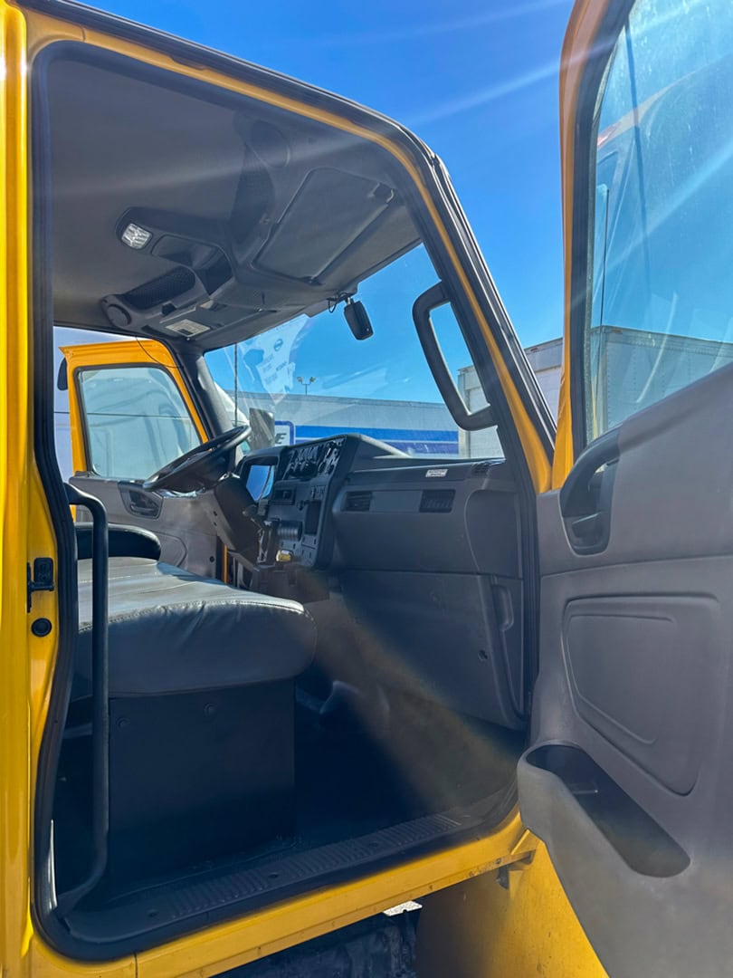 Open door view of a yellow truck’s cab under bright sunlight, showing the driver’s seat, steering wheel, dashboard, and part of the passenger seat. The sky is clear and blue outside.