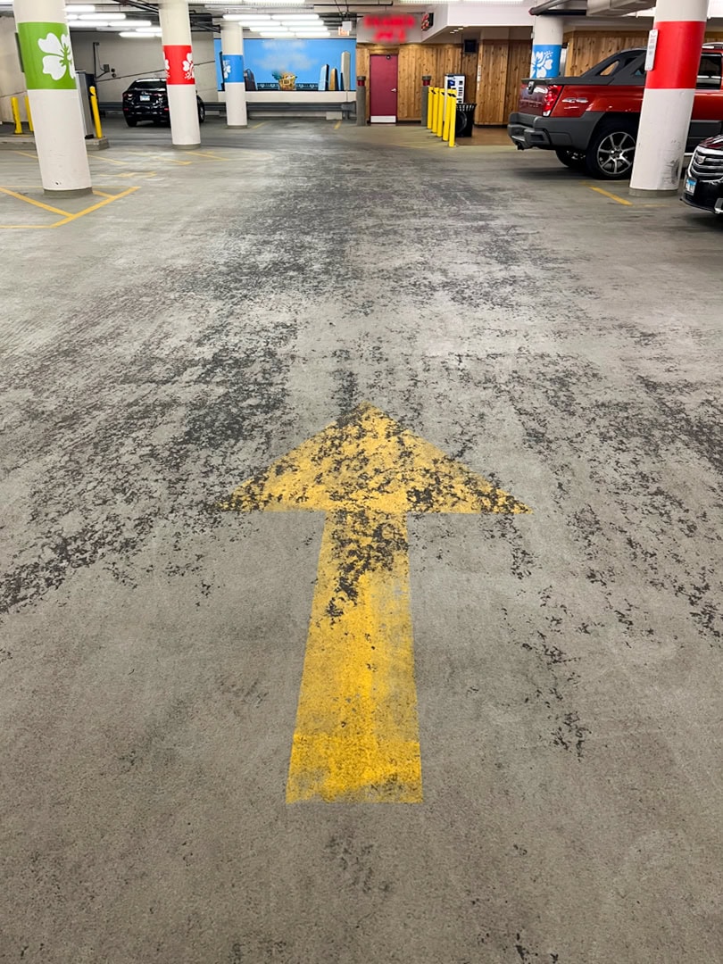 A yellow arrow is painted on the concrete floor of an indoor parking garage, pointing straight ahead. Cars are parked on the right side, and colored signs and pillars are visible in the background.
